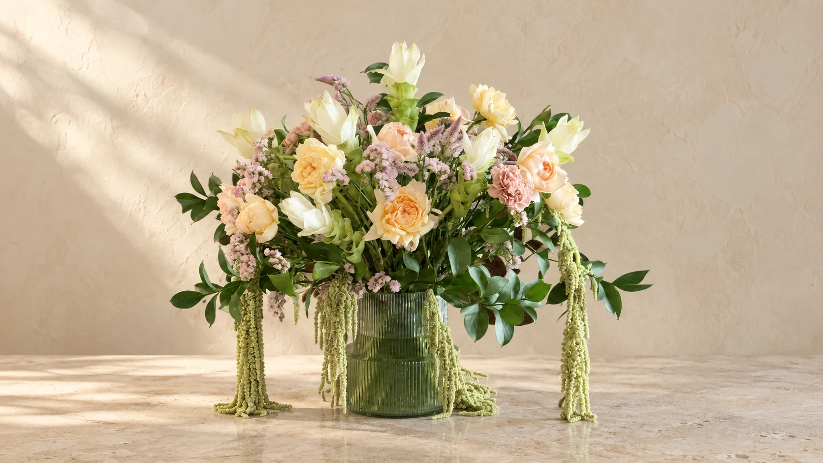 Peach roses, ivory flowers, and trailing green amaranth in a ribbed green vase, set on pale stone in soft studio light.